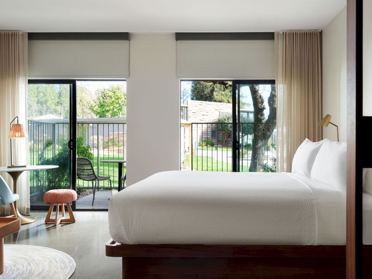 A modern bedroom with a large bed, two windows with sheer curtains, a small desk, a chair, and a view of a balcony with outdoor greenery visible.